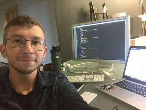 Man with glasses coding at desk with dual monitors displaying programming code and laptop in home office setup