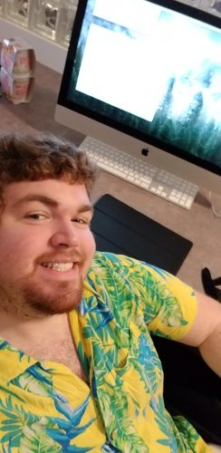 Young man in tropical yellow shirt smiling while taking selfie at computer desk with iMac and graphics tablet