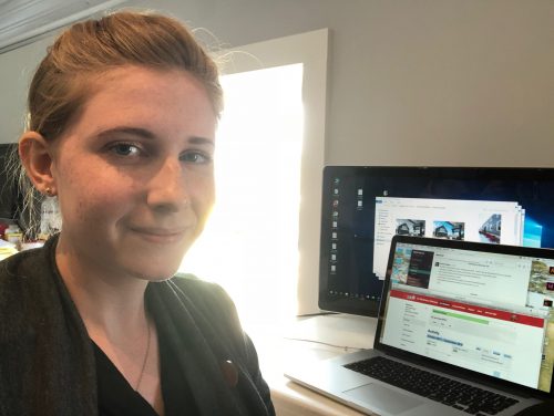 Woman working on laptop with multiple browser windows open, sitting at desk near bright window in home office setup.