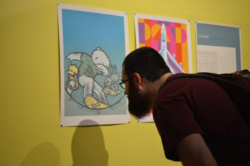 Man with glasses and beard examining colorful art prints displayed on bright yellow gallery wall