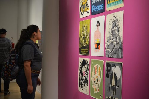 Woman viewing colorful poster exhibition on pink gallery wall displaying various artistic designs and messages.
