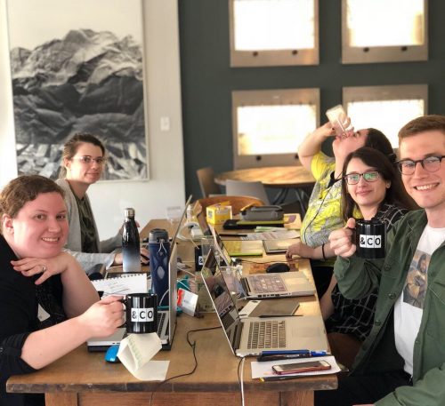 Team of five young professionals working together at wooden table with laptops and coffee mugs in modern office