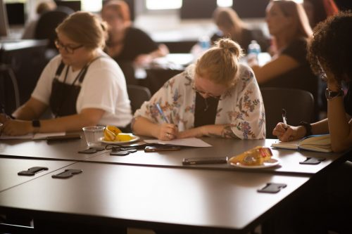Students writing and studying together at desks in a classroom workshop setting with art supplies and snacks