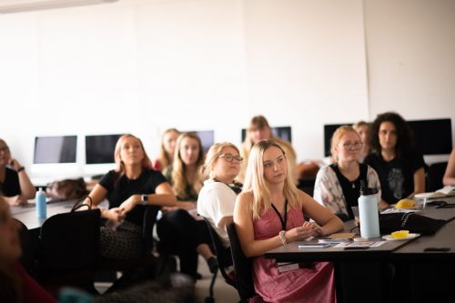 Students attentively listening during a university lecture or seminar in a modern classroom with computers and desks.