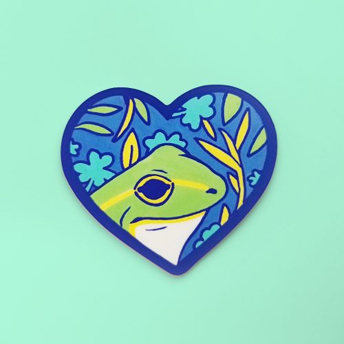 Heart-shaped enamel pin featuring green frog with plants and leaves in blue background nature design