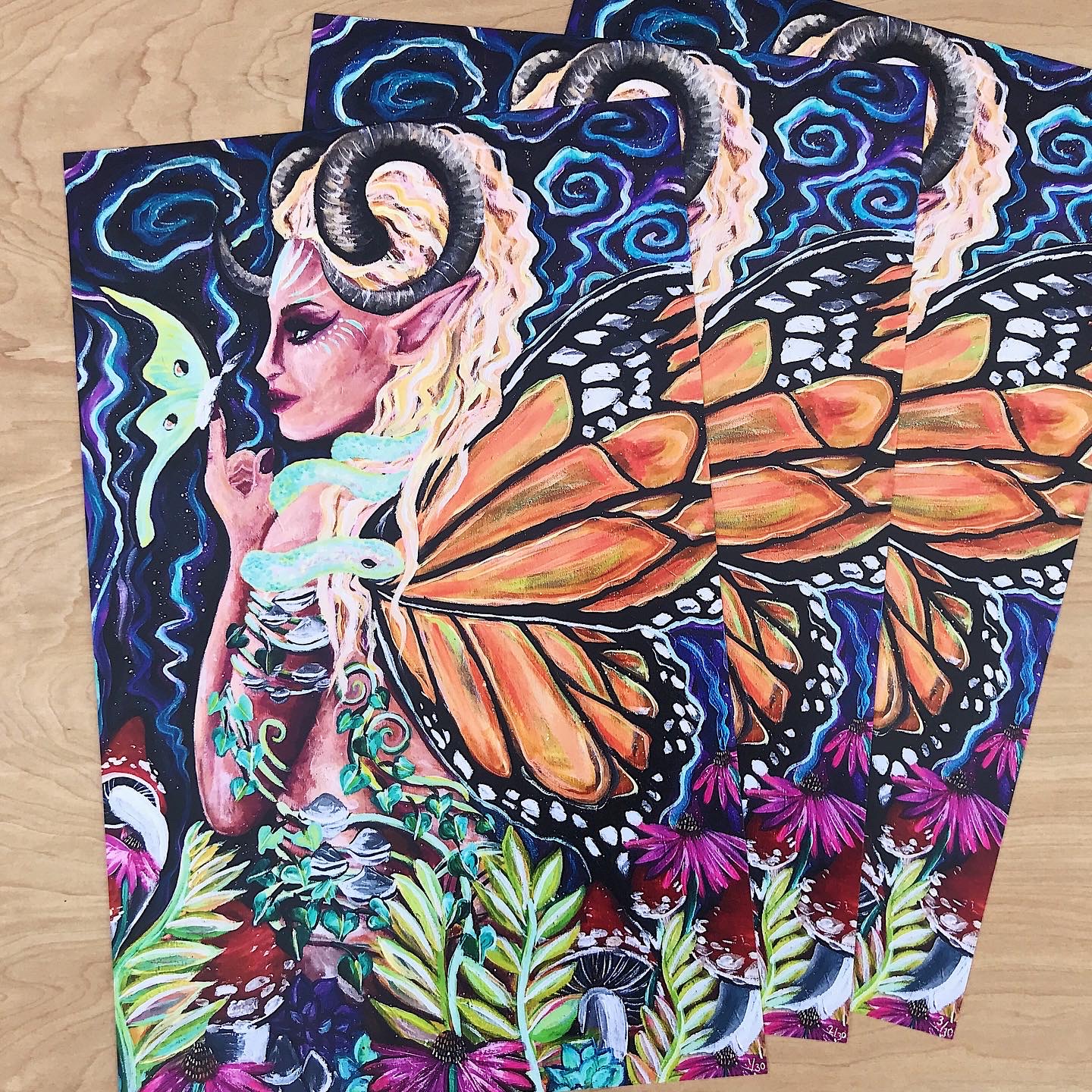Vibrant fantasy art prints featuring woman with horns, butterfly wings, and colorful flowers on wooden surface