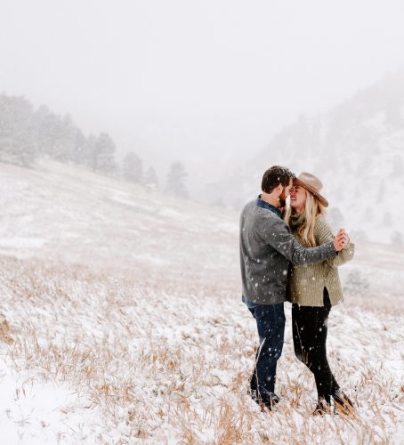 Couple embracing in snowy field during winter engagement photo session with mountains in background