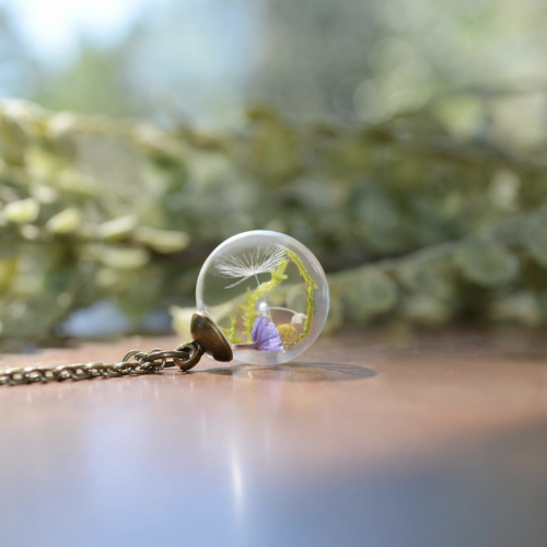 Glass sphere pendant necklace with preserved dandelion seed, moss and purple flower on wooden surface