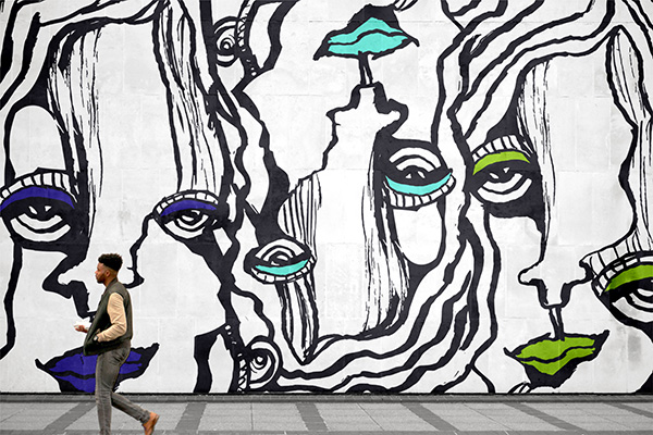 Woman walking past large black and white mural featuring abstract faces with colorful blue and green accents on white wall