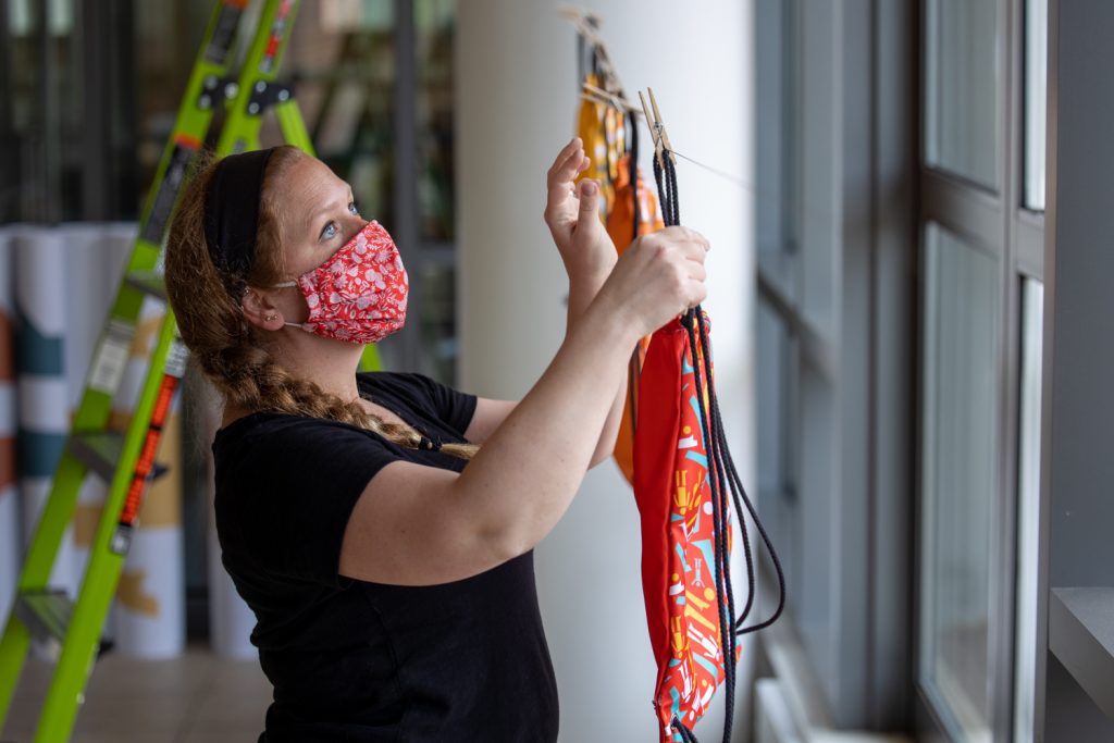 Woman wearing red floral mask hanging colorful artwork with clothespins in bright indoor studio space