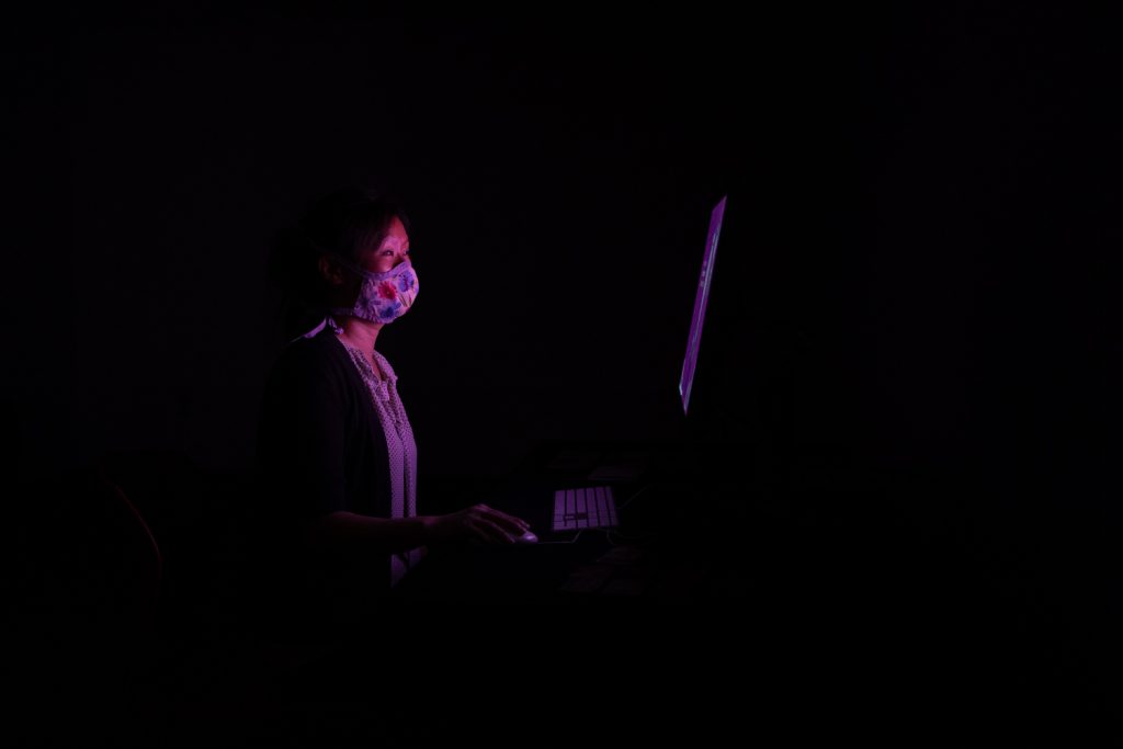 Person wearing face mask working on laptop computer in dark room with purple lighting