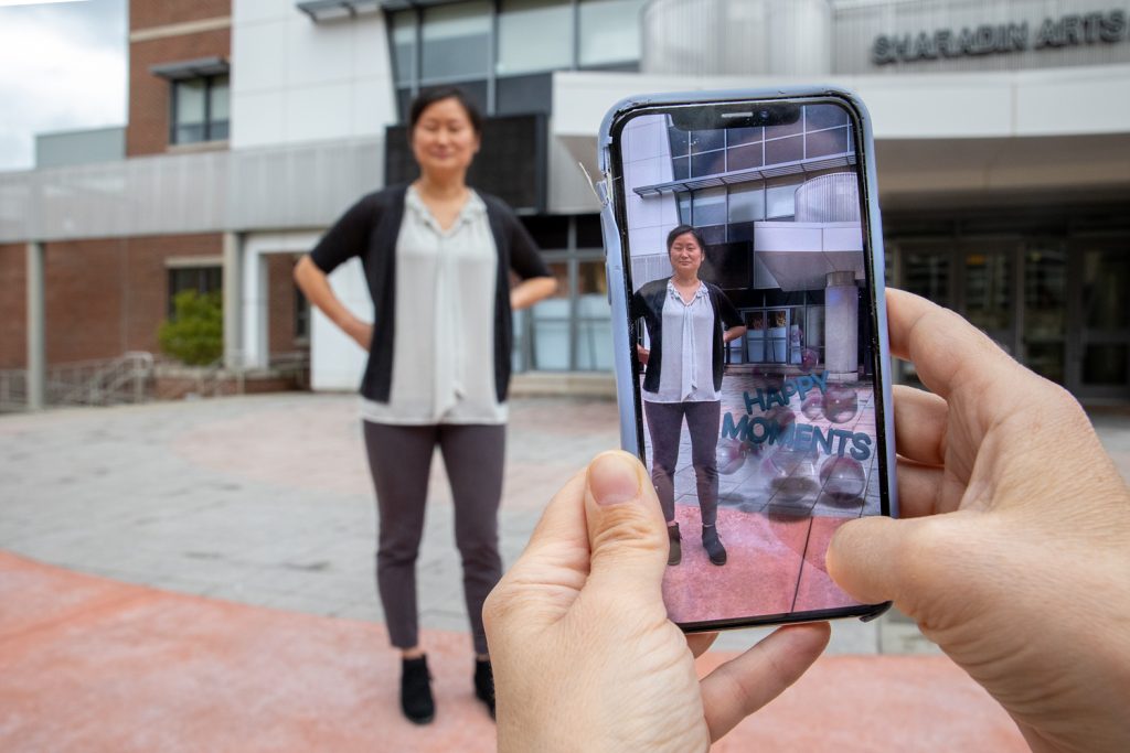 Woman using smartphone AR app with Happy Moments text overlay while photographing person outside Sharadin Arts building