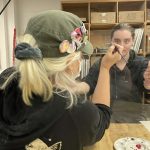 Person in green hat with pins applying face paint while looking in mirror in art classroom with storage shelves