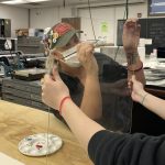 Student painting on clear acrylic sheet in art classroom with paint palette and protective barrier during COVID-19