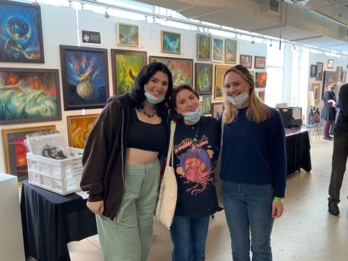 Three young women wearing face masks posing together at an art gallery exhibition with colorful paintings on walls