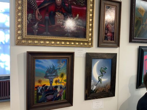 Art gallery wall displaying framed paintings including surreal deer artwork and frog on crescent moon piece