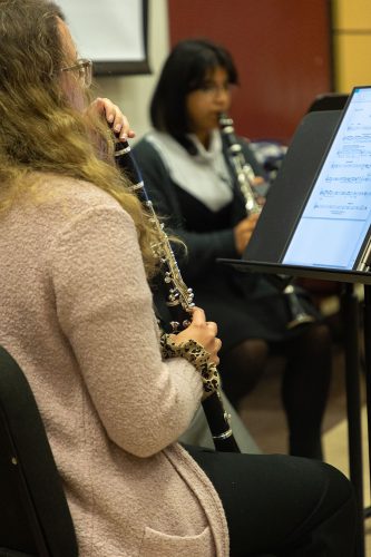 Woman playing clarinet with sheet music while another musician performs in background during indoor concert or rehearsal