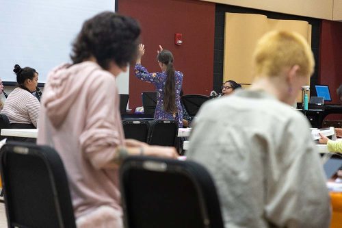 Woman in blue floral dress presenting to students in modern classroom with laptops and tiered seating