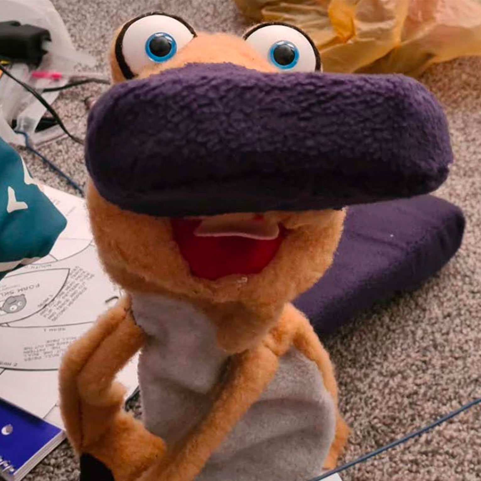 Colorful plush toy character with googly eyes, purple knit hat, orange body and open red mouth sitting on carpet
