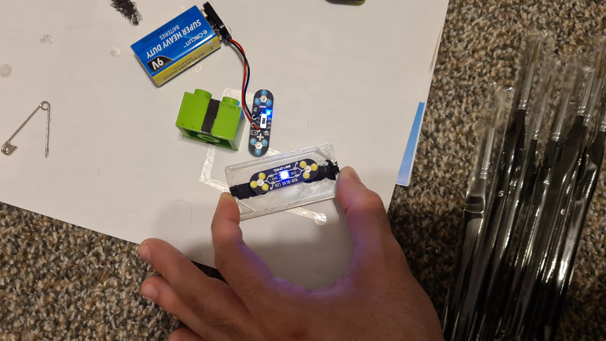 Person holding 3D printed electronics project with circuit board, 9V battery, and LED components on white surface
