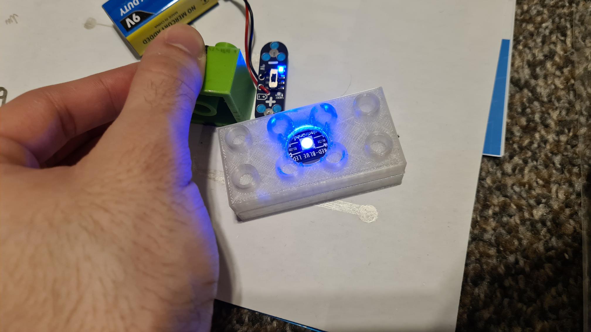 Hand holding UV light over 3D printed case with glowing blue circuit board and electronic components inside